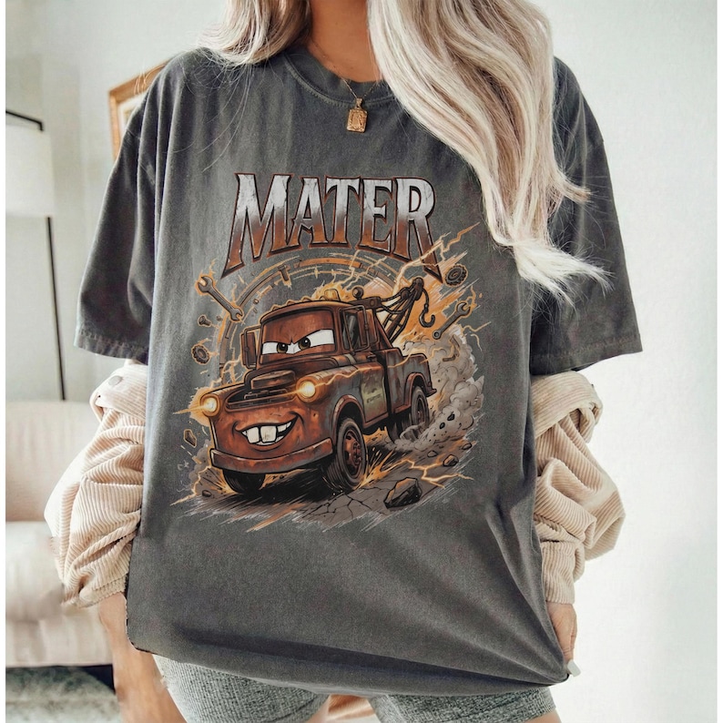 Pu&ograve; includere: T-shirt grigio scuro con un'immagine a cartoni animati di Mater del film Cars. La grafica include la parola "MATER" in arancione e marrone, con fulmini e strumenti che circondano il personaggio. La maglietta &egrave; uno stile unisex.