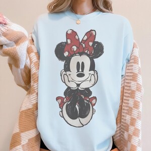 May include: Light blue sweatshirt featuring a distressed graphic of Minnie Mouse. Minnie is depicted with a red polka dot bow, black ears, and a red skirt. The sweatshirt is a casual, comfortable style.