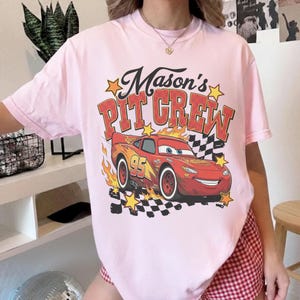 May include: A light pink t-shirt featuring a graphic of a red race car with the text "Mason's PIT CREW". The design includes flames, stars, and a checkered flag, creating a racing theme.