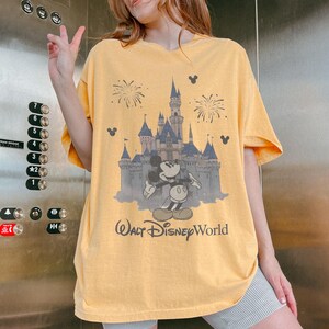 Puede incluir: Camiseta amarilla con un dibujo de Mickey Mouse frente a un castillo y fuegos artificiales, con el texto "Walt Disney World". La camiseta tiene mangas cortas y un corte relajado. El dibujo es en tonos grises y azules.