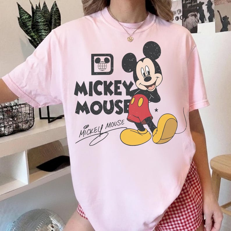 May include: A light pink t-shirt featuring a graphic of Mickey Mouse. The design includes the words "Mickey Mouse" in black, along with an image of Mickey Mouse in his classic outfit. The shirt has short sleeves.