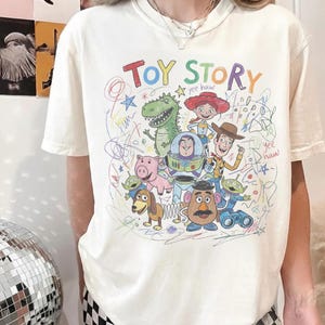 May include: Cream-colored t-shirt featuring a colorful, hand-drawn illustration of characters from the animated film "Toy Story." The design includes the words "Toy Story" in rainbow colors and various characters like Woody, Buzz Lightyear, and Rex.
