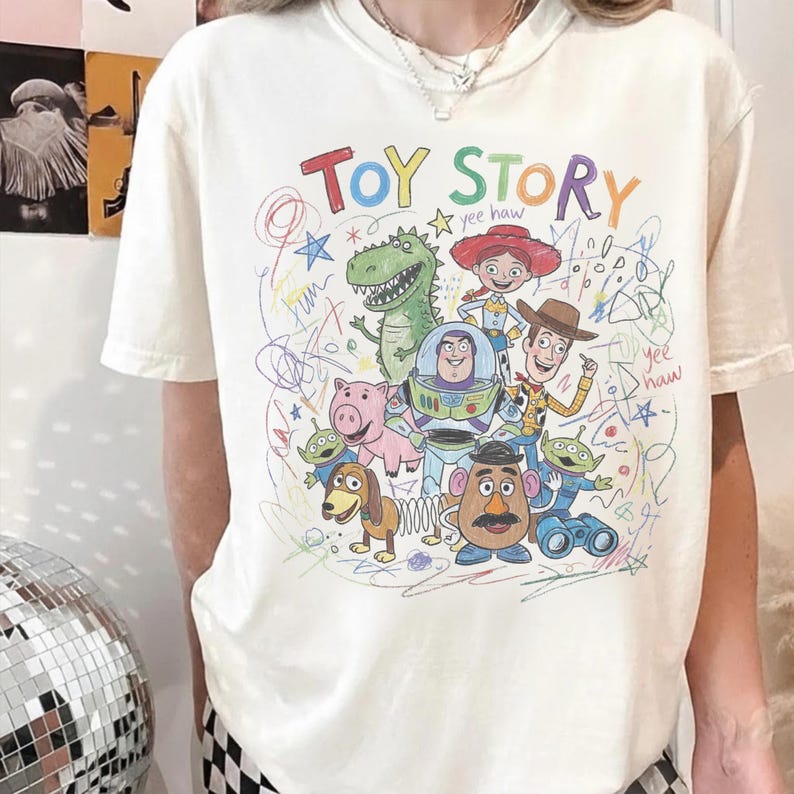 May include: Cream-colored t-shirt featuring a colorful, hand-drawn illustration of characters from the animated film "Toy Story." The design includes the words "Toy Story" in rainbow colors and various characters like Woody, Buzz Lightyear, and Rex.