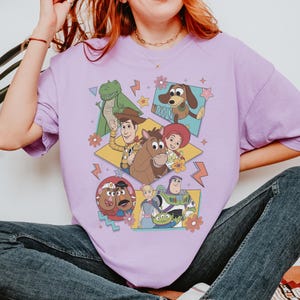 Pode incluir: Uma camiseta lil&aacute;s com um gr&aacute;fico colorido dos personagens de Toy Story. O design apresenta Woody, Buzz Lightyear, Jessie, Rex, Slinky Dog e Sr. e Sra. Cabe&ccedil;a de Batata, com detalhes florais e raios.