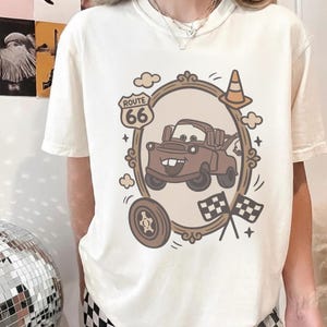 May include: Cream-colored t-shirt featuring a cartoon image of Mater from the movie Cars, framed by a decorative border with Route 66 signage, clouds, and checkered flags. The design includes a traffic cone and a tire.
