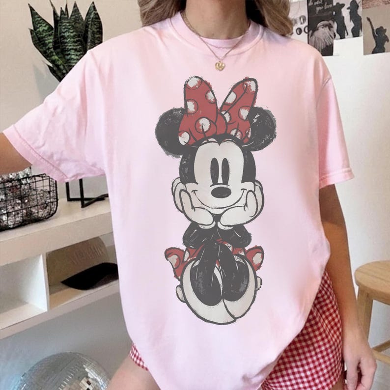 May include: A light pink t-shirt featuring a graphic of Minnie Mouse. The cartoon character is depicted in black and white with a red polka dot bow. The shirt is a casual, oversized fit.