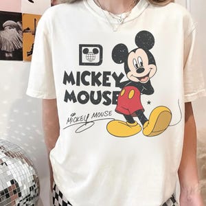 May include: Cream-colored t-shirt featuring a graphic of Mickey Mouse. The design includes the words "Mickey Mouse" in bold, black letters, with a stylized signature below. Mickey Mouse is depicted in his classic outfit.