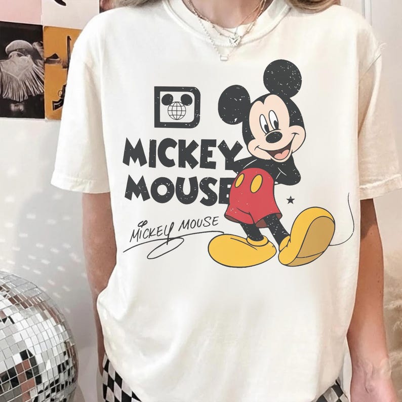 May include: Cream-colored t-shirt featuring a graphic of Mickey Mouse. The design includes the words "Mickey Mouse" in bold, black letters, with a stylized signature below. Mickey Mouse is depicted in his classic outfit.