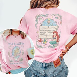 May include: Pale pink t-shirt featuring a mermaid design, musical notes, and the text "Wish I could be Part of That World." The back of the shirt has a mermaid, musical notes, and sea creatures. The t-shirt is a light pink colour.