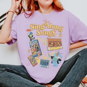Puede incluir: Camiseta lila con la inscripci&oacute;n "Disney Sing Along Songs" en amarillo. La camiseta presenta gr&aacute;ficos de personajes de dibujos animados y cintas de cassette. Dise&ntilde;o de inspiraci&oacute;n vintage.