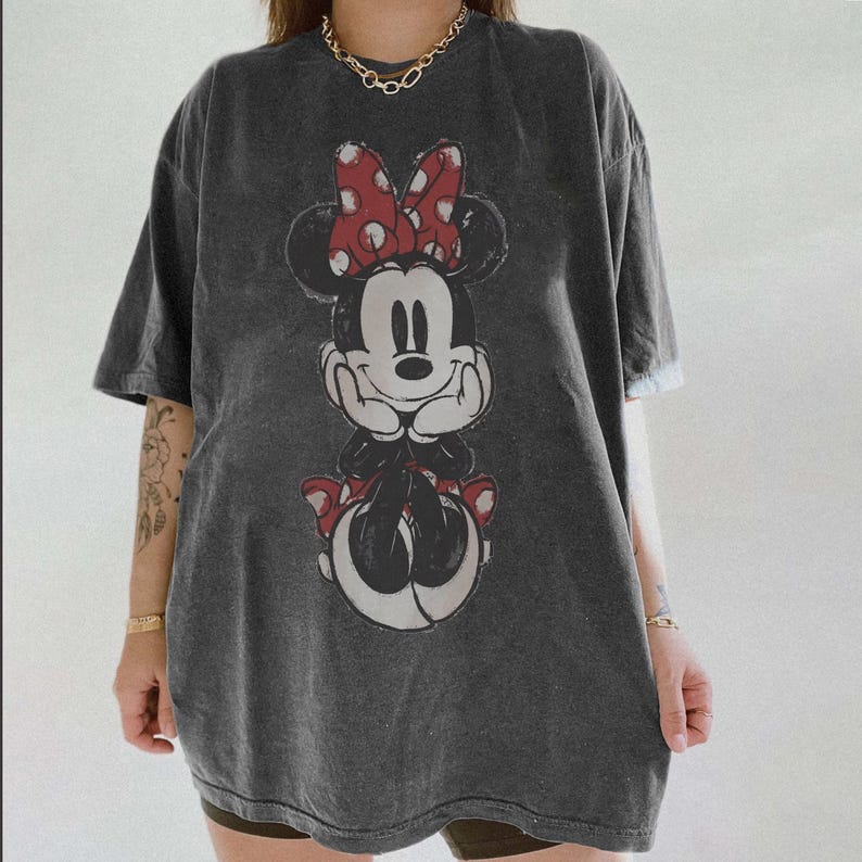 May include: Dark gray t-shirt featuring a large graphic of Minnie Mouse. Minnie is depicted in black and white with a red polka dot bow and red shoes. The shirt has short sleeves and a relaxed fit, suitable for casual wear.