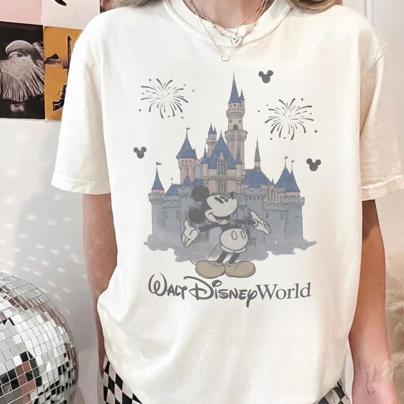Puede incluir: Camiseta color crema con un gr&aacute;fico del castillo de Cenicienta, Mickey Mouse, fuegos artificiales y el texto "Walt Disney World". El dise&ntilde;o es en tonos azules, grises y negros.