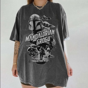 以下が含まれることがあります： スターウォーズシリーズ「マンダロリアンとグローグ」のグラフィックが特徴の濃いグレーのTシャツ。デザインにはマンダロリアンのヘルメット、グローグ、「This is the Way」というフレーズが含まれています。シャツはヴィンテージ感があります。