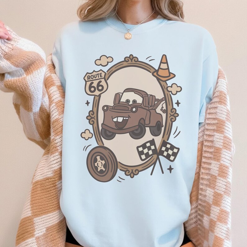 May include: Light blue sweatshirt featuring a cartoon illustration of a brown tow truck inside a decorative frame. The design includes Route 66 signage, a traffic cone, checkered flags, and a tire. The sweatshirt is a crew neck style.