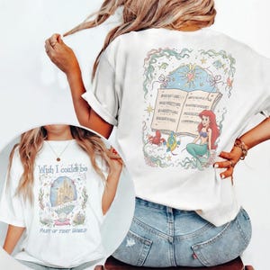 May include: White t-shirt with a colourful illustration of a mermaid with red hair, sitting in front of an open music book. The back of the shirt has the text "Wish I could be part of that world". The design includes sea creatures and a castle.