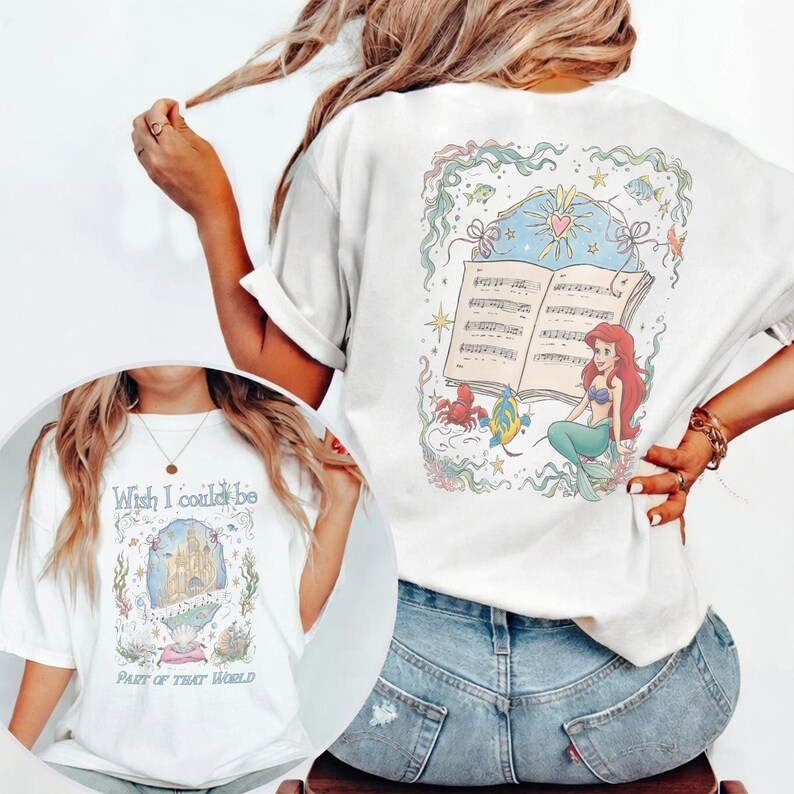 May include: White t-shirt with a colourful illustration of a mermaid with red hair, sitting in front of an open music book. The back of the shirt has the text "Wish I could be part of that world". The design includes sea creatures and a castle.