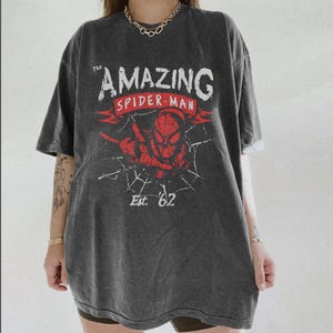 May include: Dark grey t-shirt with a distressed Spider-Man graphic. The design features the words "The Amazing Spider-Man" in white and red, and "Est. '62". The shirt has short sleeves and a loose fit, suitable for casual wear.