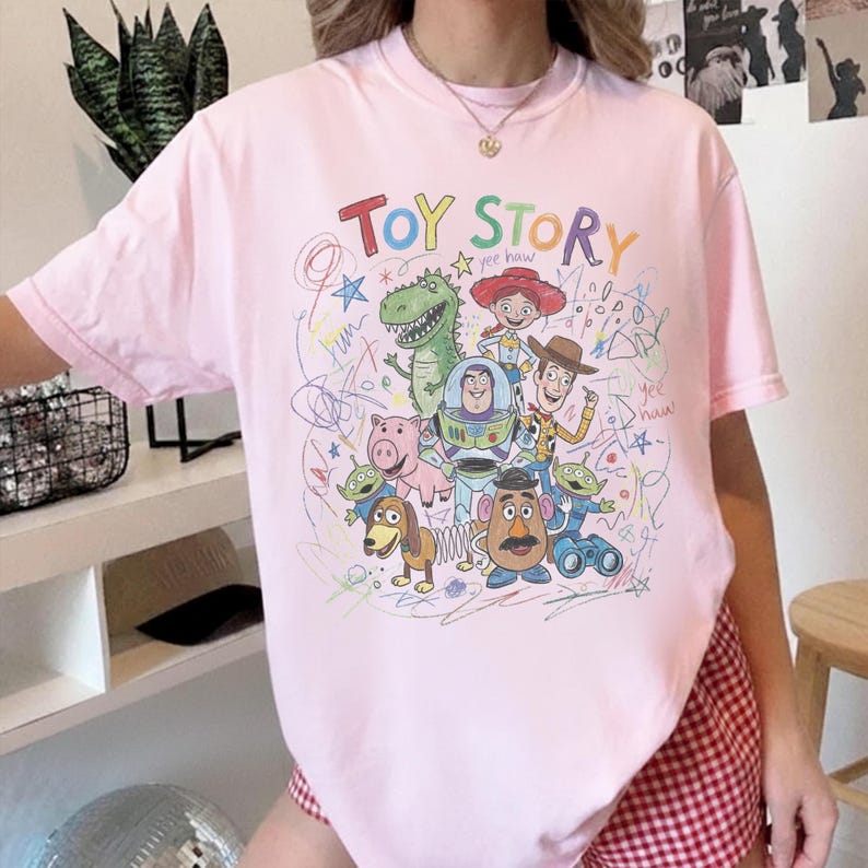 May include: A light pink t-shirt featuring the words "TOY STORY" in colorful letters, with a graphic of characters from the movie. The design includes a green dinosaur, Woody, Buzz Lightyear, and other characters.