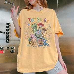 May include: Yellow short-sleeved t-shirt featuring the text "Toy Story" above a colorful graphic of characters from the animated film. The design includes Woody, Buzz Lightyear, Rex, and other characters. The shirt has a relaxed fit.