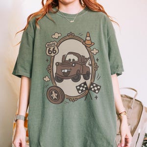 May include: Green t-shirt featuring a cartoon image of Mater from the movie Cars, framed by a decorative oval with Route 66 text. The design includes clouds, a traffic cone, a tire, and checkered flags. The shirt is a comfortable, casual style.