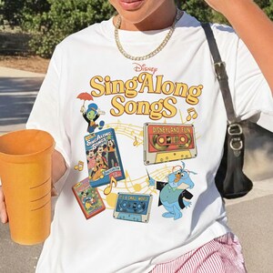 Puede incluir: Camiseta blanca con el logo de Disney "Sing Along Songs" en amarillo y naranja. Presenta personajes de dibujos animados de Disney y cintas de cassette. Una persona sostiene una taza grande de color naranja.