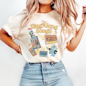 Puede incluir: Camiseta color crema con el logo de Disney "Sing Along Songs" y varios personajes de dibujos animados y cintas de cassette. La camiseta tiene una est&eacute;tica vintage, con un aspecto ligeramente descolorido. Tambi&eacute;n se ve el texto "Disneyland Fun".