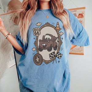 May include: Light blue t-shirt featuring a brown cartoon tow truck inside an oval frame with the text "Route 66". The design includes a traffic cone, checkered flags, and a tire.