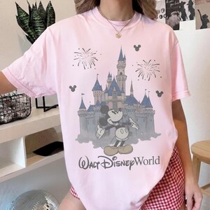 Puede incluir: Camiseta rosa claro con un dise&ntilde;o de Mickey Mouse frente a un castillo, fuegos artificiales y el texto "Walt Disney World". El dise&ntilde;o es en azul, gris y negro.