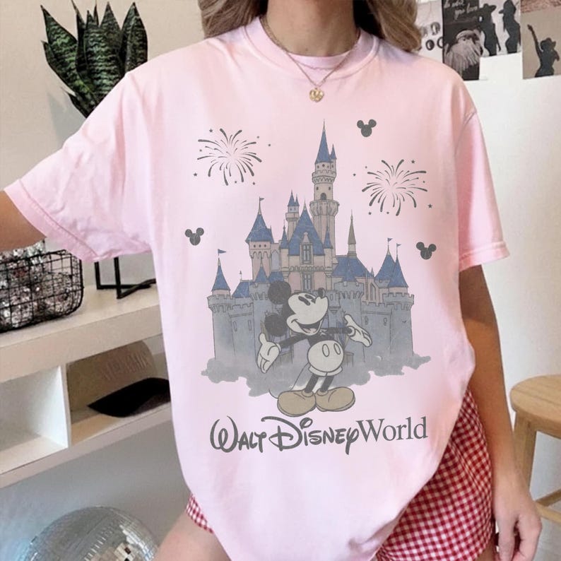 Puede incluir: Camiseta rosa claro con un dise&ntilde;o de Mickey Mouse frente a un castillo, fuegos artificiales y el texto "Walt Disney World". El dise&ntilde;o es en azul, gris y negro.