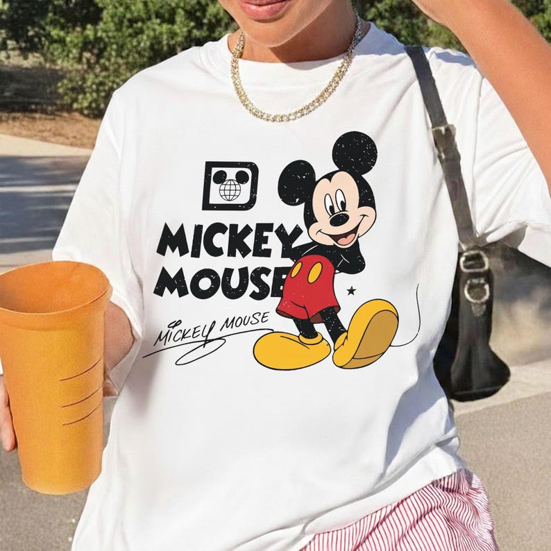 May include: White t-shirt featuring a graphic of Mickey Mouse. The design includes the words "Mickey Mouse" in a bold, black font, and a drawing of Mickey Mouse in his classic outfit. The shirt also has a small graphic of a globe.