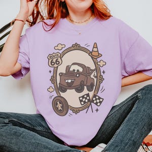 May include: A light purple t-shirt featuring a cartoon image of Mater from the movie Cars, framed by a decorative border with Route 66 signage. Additional elements include a tire, clouds, and checkered flags.