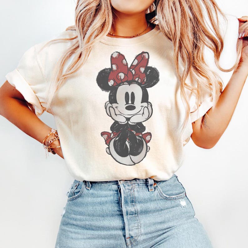 May include: Cream-colored t-shirt featuring a seated Minnie Mouse graphic. Minnie Mouse is depicted in black and white with a red polka-dot bow and accents. The graphic is centered on the shirt, which has rolled-up sleeves.