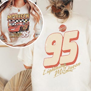 Op de afbeelding: Cr&egrave;mekleurig T-shirt met een Lightning McQueen-afbeelding en het nummer 95. De voorkant van het shirt toont Lightning McQueen met de tekst "Lightning McQueen", "Think Fast" en "95". De achterkant heeft een grote "95" en de tekst "Lightning McQueen".