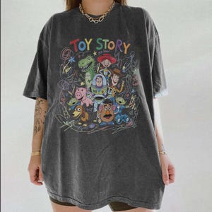 May include: Dark gray t-shirt featuring a colorful graphic of characters from the animated film "Toy Story." The design includes the text "Toy Story" in a rainbow font, along with images of Woody, Buzz Lightyear, and other characters.