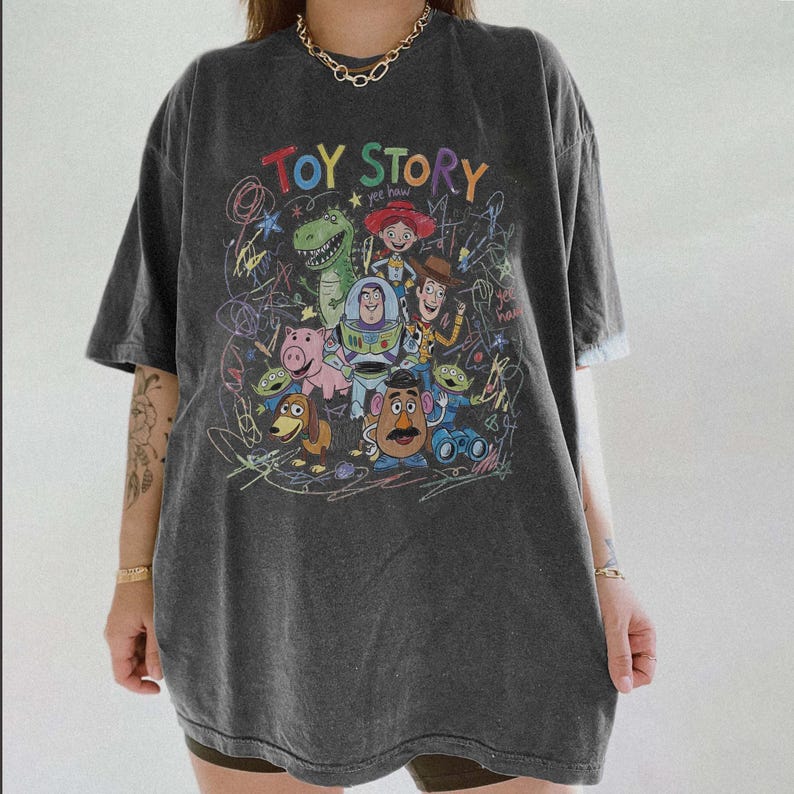 May include: Dark gray t-shirt featuring a colorful graphic of characters from the animated film "Toy Story." The design includes the text "Toy Story" in a rainbow font, along with images of Woody, Buzz Lightyear, and other characters.
