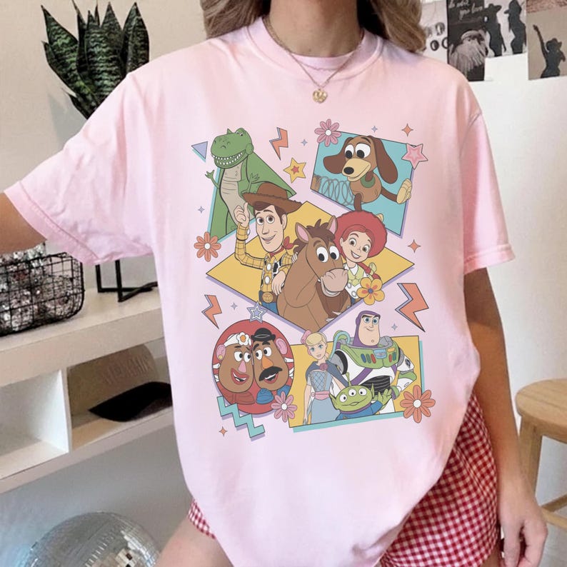 Pode incluir: Uma camiseta rosa claro com um gr&aacute;fico colorido de v&aacute;rios personagens do filme de anima&ccedil;&atilde;o "Toy Story". O design inclui imagens de Woody, Buzz Lightyear, Rex e outros personagens, em um fundo geom&eacute;trico com flores e raios.