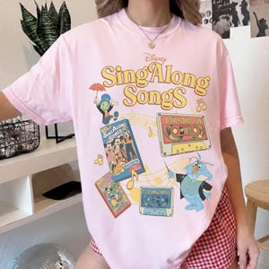 Puede incluir: Camiseta rosa claro con el logotipo de Disney Sing Along Songs y varios personajes de dibujos animados y cintas de cassette. El dise&ntilde;o incluye el texto "Disneyland Fun" y "It's a Small World". La camiseta tiene cuello redondo y mangas cortas.