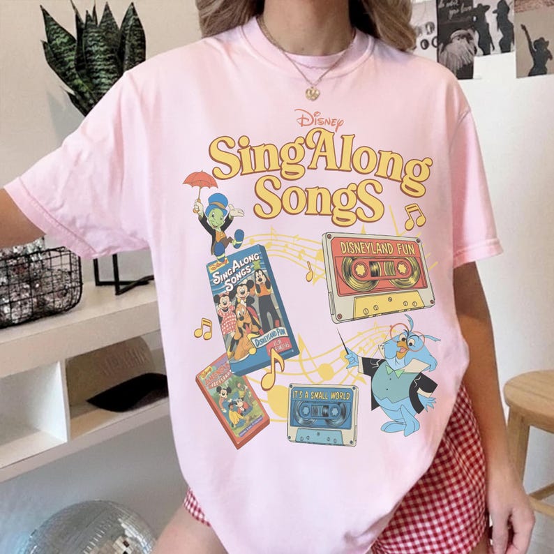 Puede incluir: Camiseta rosa claro con el logotipo de Disney Sing Along Songs y varios personajes de dibujos animados y cintas de cassette. El dise&ntilde;o incluye el texto "Disneyland Fun" y "It's a Small World". La camiseta tiene cuello redondo y mangas cortas.