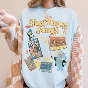 Puede incluir: Camiseta azul claro con un dise&ntilde;o de Disney "Sing Along Songs". La camiseta presenta personajes de dibujos animados y cintas de cassette. El texto "Disneyland Fun" y "It's a Small World" es visible en las cintas.