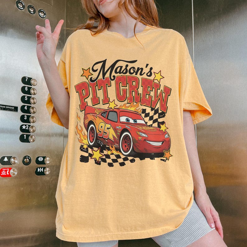 May include: Mustard yellow t-shirt with a graphic of Lightning McQueen from the movie Cars. The text "Mason's PIT CREW" is printed above the car in red and black. The shirt has a vintage, worn look.