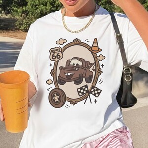 May include: White t-shirt featuring a cartoon image of a brown tow truck inside a decorative frame, with Route 66 text. The design includes a tire, clouds, checkered flags, and a traffic cone. The shirt is a casual wear item.