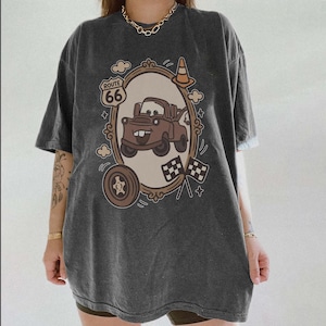 May include: Dark gray t-shirt featuring a cartoon image of a brown tow truck inside an ornate frame. The design includes the Route 66 logo, a tire, checkered flags, and a traffic cone. The shirt has short sleeves and a relaxed fit.