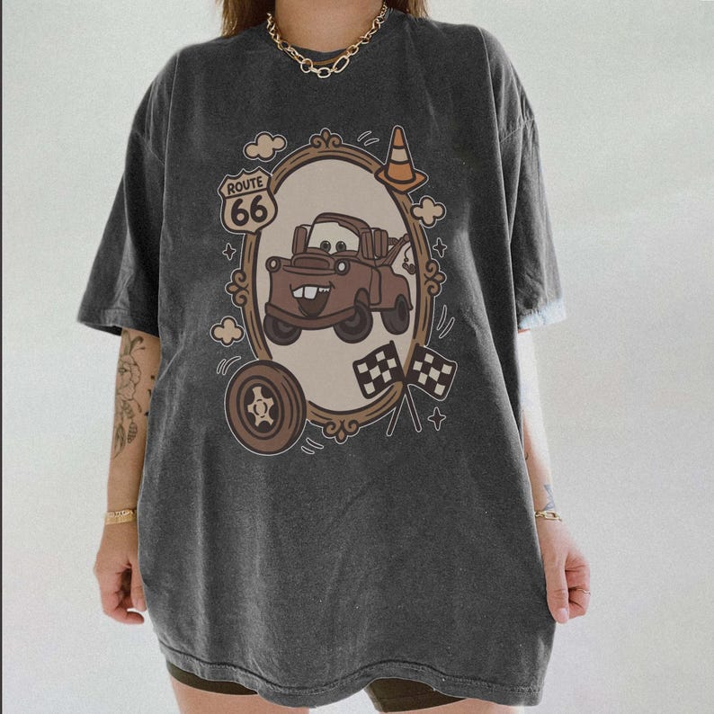 May include: Dark gray t-shirt featuring a cartoon image of a brown tow truck inside an ornate frame. The design includes the Route 66 logo, a tire, checkered flags, and a traffic cone. The shirt has short sleeves and a relaxed fit.