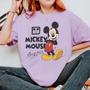 May include: A lilac-colored t-shirt featuring a graphic of Mickey Mouse. The design includes Mickey in his classic pose, with red shorts, yellow shoes, and black gloves. The text "Mickey Mouse" is printed above the character in a distressed font.