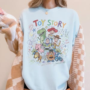 May include: Light blue sweatshirt featuring a colorful graphic of characters from the animated film "Toy Story." The design includes the words "Toy Story" in a playful font, along with drawings of Woody, Buzz Lightyear, and other characters.