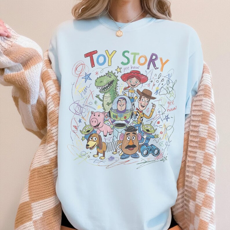May include: Light blue sweatshirt featuring a colorful graphic of characters from the animated film "Toy Story." The design includes the words "Toy Story" in a playful font, along with drawings of Woody, Buzz Lightyear, and other characters.
