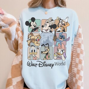 May include: Light blue sweatshirt featuring a grid of Disney characters including Mickey Mouse, Minnie Mouse, Goofy, Chip and Dale, Stitch, Donald Duck, Pluto, and Daisy Duck. The words "Walt Disney World" are printed in black script below the grid.