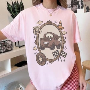 May include: Light pink t-shirt featuring a cartoon car design within a decorative frame, with a Route 66 sign, a traffic cone, and checkered flags. The design is in shades of brown, beige, and white. The shirt is a casual wear item.