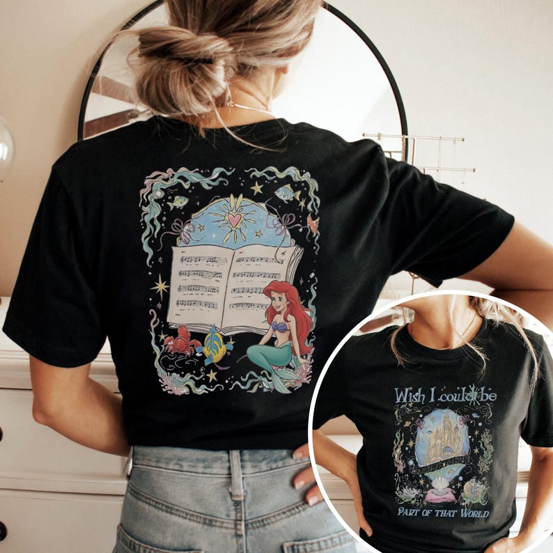 May include: Black t-shirt with a vibrant illustration of Ariel from The Little Mermaid. The design includes sheet music, a heart, stars, and sea creatures. The shirt also has the text "Wish I could be part of that world."
