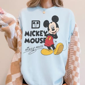 May include: Light blue sweatshirt featuring a graphic of Mickey Mouse. The design includes Mickey in his classic outfit, with the words "Mickey Mouse" in bold letters. A small graphic of a globe is also present.